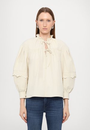 Blouse couleur crème avec un motif texturé, col volanté, fermeture à nouer, et manches bouffantes avec des bords festonnés. Associée à un jean bleu.