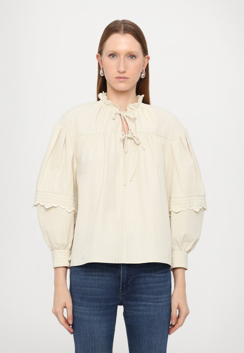 Cream-colored blouse with a textured pattern, ruffled collar, tie closure, and puffed sleeves with scalloped edges. Paired with blue jeans.