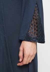 Donkerblauwe stoffen jurk met uitlopende mouwen. Kantdetail aan de mouwranden met een geometrisch patroon. Zachte textuur met een gladde afwerking.