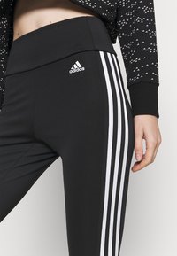 Svarta träningsleggings med hög midja, som har tre vita ränder längs varje sida och Adidas-logotypen fram. Stretchigt material.