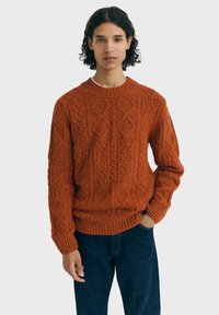 Personne avec des cheveux bouclés et fonc�és, portant un pull en tricot texturé de couleur rouille et un jean bleu foncé, se tenant avec une main dans la poche.
