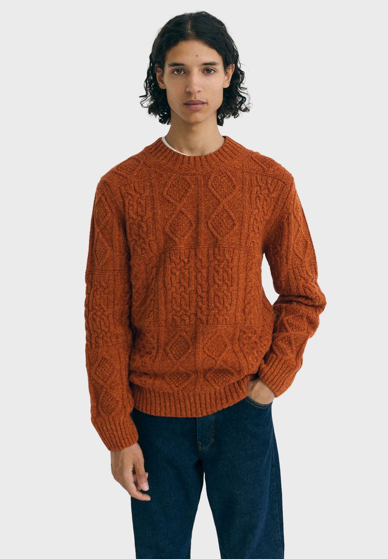 Personne avec des cheveux bouclés et foncés, portant un pull en tricot texturé de couleur rouille et un jean bleu foncé, se tenant avec une main dans la poche.