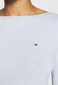Lähikuva henkilöstä, jolla on päällään vaaleansininen paita, jossa on pieni suorakulmainen logo tummansinisellä, valkoisella ja punaisella rinnassa.