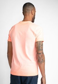 Majica s kratkimi rokavi v svetli koralni barvi, izdelana iz mehkega materiala. Ima okrogel izrez in običajen kroj. Vidna tetovaža na podlahti.