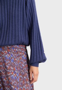 Marine-blauer Rippstrickpullover mit Glockenärmel, der eine glatte Textur aufweist. Kombiniert mit einem gemusterten Rock in Lila-, Rot- und Weißtönen.