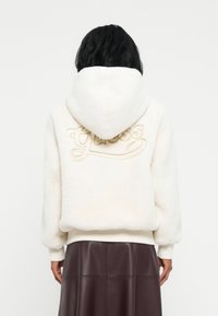 Persona con capelli neri che indossa un hoodie bianco e peloso con la scritta "Guess" ricamata sul retro e una gonna di pelle marrone scuro.