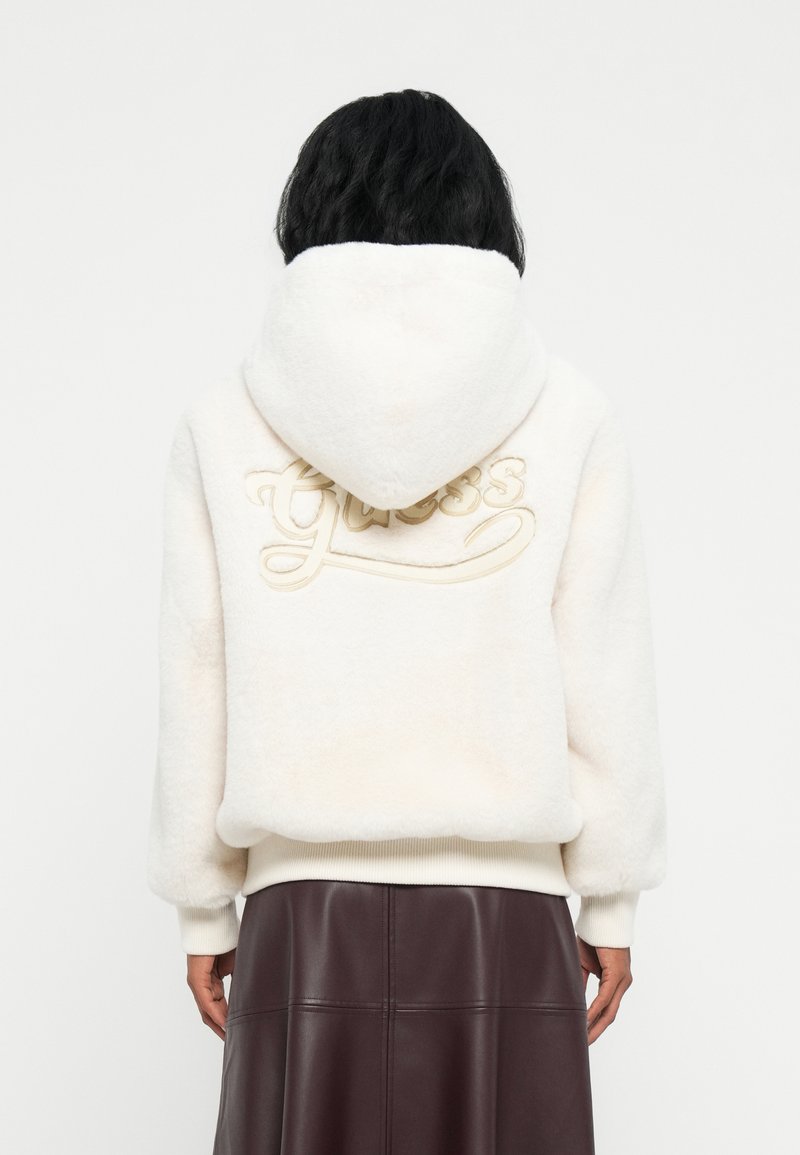 Persona con capelli neri che indossa un hoodie bianco e peloso con la scritta "Guess" ricamata sul retro e una gonna di pelle marrone scuro.