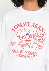 Felpa bianca con una grafica rossa di un cuoco che tiene una pizza, con il testo "TOMMY JEANS NEW YORK PIZZERIA". Texture morbida, vestibilità comoda.