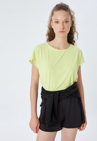 Limonengrünes Kurzarmoberteil, kombiniert mit schwarzen Shorts, mit lockerer Passform und einem Bund, der mit einem schwarzen Stoff gebunden ist. Einfarbiger Hintergrund.