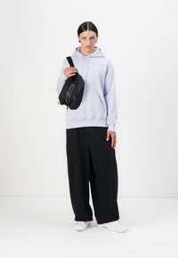 Hoodie lilás claro com bolso frontal, combinado com calças pretas largas e uma pequena bolsa cruzada preta com fecho. Ténis brancos.