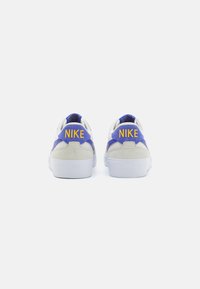 Tenisi Nike în gri, mov și alb. Prezintă materiale din daim și plasă. Partea din spate include logo-ul "NIKE" de culoare galbenă pe accente mov.