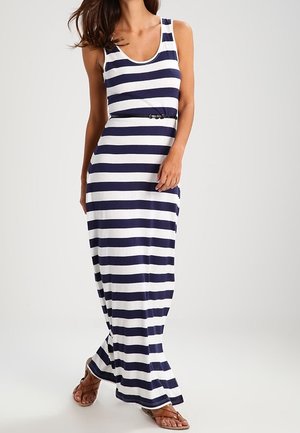 Mujer que lleva un vestido maxi sin mangas con rayas horizontales azul marino y blancas, con un cinturón negro y sandalias marrones, de pie sobre un fondo liso.
