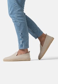 Espadrillas di un beige chiaro con tomaia in tessuto testurizzato, suola in corda di juta e tagli laterali, indossate con jeans a sigaretta azzurri chiari.