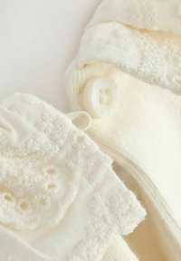 Primo piano di un tessuto color crema con ricami floreali, trame di maglieria e un bottone bianco tondo cucito.