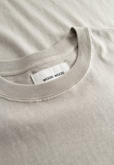 Gros plan sur un T-shirt beige à col rond avec une étiquette de marque blanche visible portant l'inscription « WOOD WOOD » à l'intérieur du col.