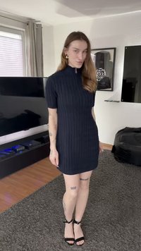 Φόρεμα σε ναυτικό μπλε ριμπ πλεκτό με κοντά μανίκια και ψηλό γιακά με φερμουάρ· συνδυασμένο με μαύρα τακούνια με λουράκια. Ανοιχτόχρωμο δέρμα, ορατά τατουάζ στα πόδια.
