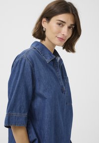 Denim srajca v temno modri barvi, z ovratnikom, kratkimi rokavi, sprednjim žepom in diskretnimi šivi. Gladka tekstura.