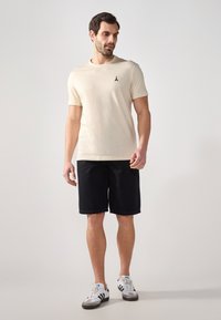 Lys beige kortermet t-skjorte med en liten svart logo, kombinert med mørkeblå shorts og hvite sneakers med svarte detaljer.