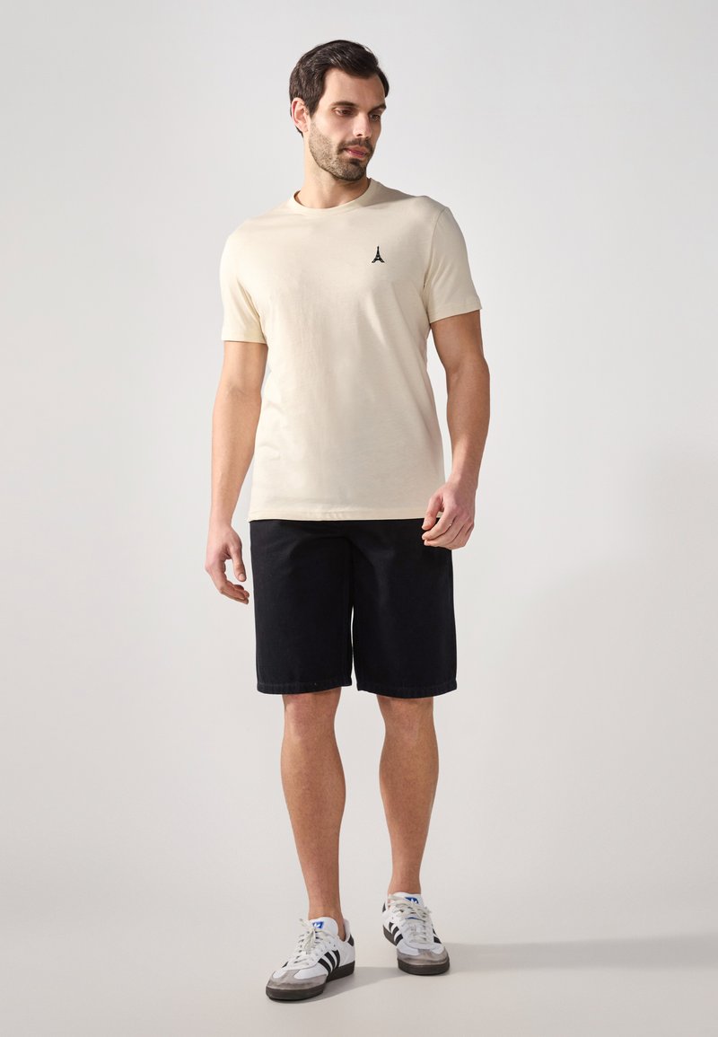 Lys beige kortermet t-skjorte med en liten svart logo, kombinert med mørkeblå shorts og hvite sneakers med svarte detaljer.