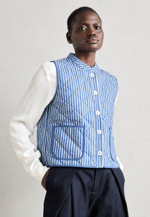 Gestepptes blau-weißes gestreiftes Weste mit Mandarin-Kragen, ausgestattet mit einer Knopfleiste und einer Seitentasche, getragen über einem gerippten weißen Shirt.