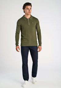 Polo in maglina verde oliva con colletto con zip, polsini e orlo a costine, abbinato a pantaloni blu navy e sneakers bianche.