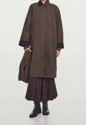 Person wearing a long brown coat with black cuffs, holding a brown knit sweater, paired with a dark pleated skirt and black ankle boots.