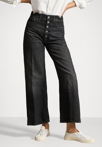 Jeans de pierna ancha negros de cintura alta con cinco botones plateados, combinados con una camisa blanca y zapatillas blancas de cordones sobre un fondo gris.