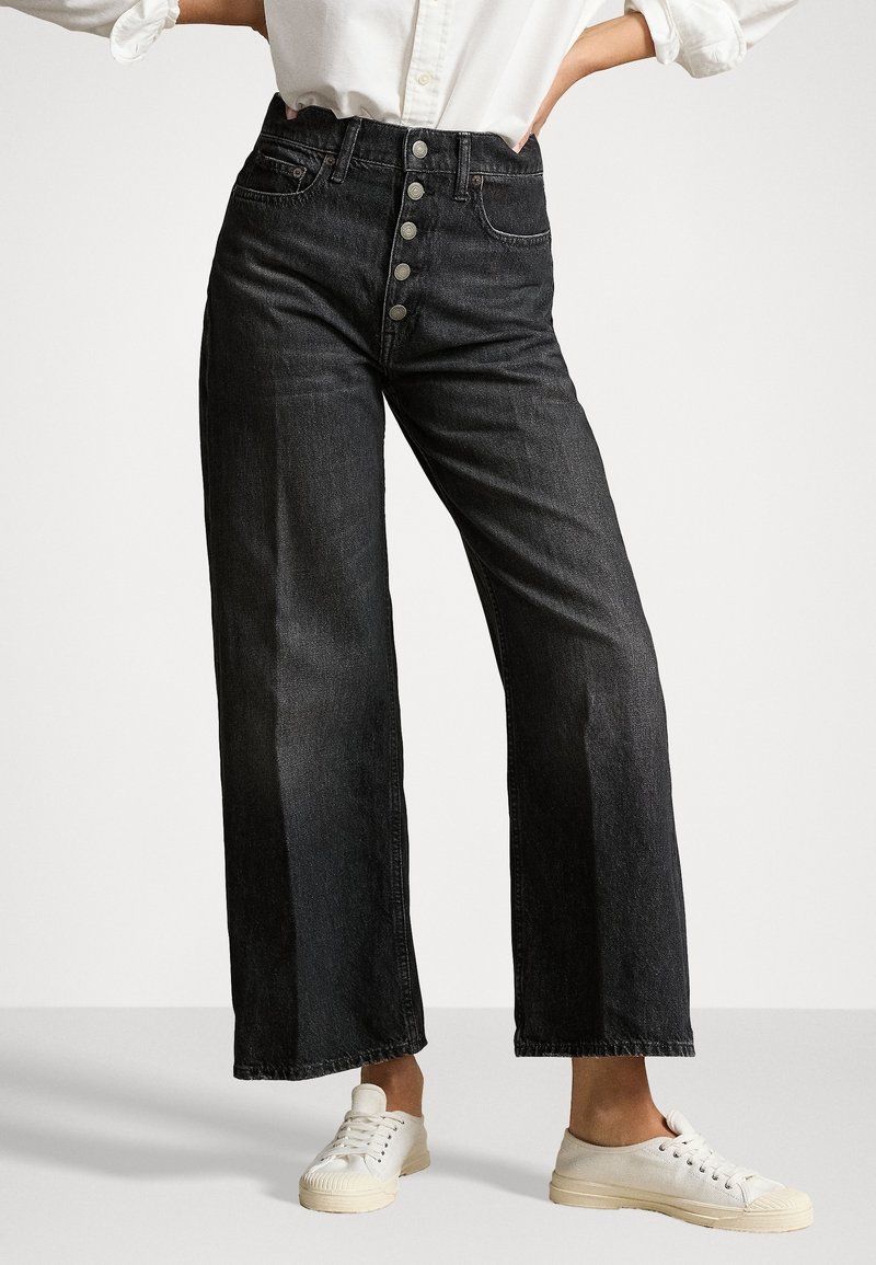 Jeans de pierna ancha negros de cintura alta con cinco botones plateados, combinados con una camisa blanca y zapatillas blancas de cordones sobre un fondo gris.