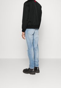 Blouson aviateur noir avec poignets côtelés, accompagné d'un jean bleu clair déchiré et de bottes noires épaisses. Étiquette rouge notable à l'arrière de la veste.