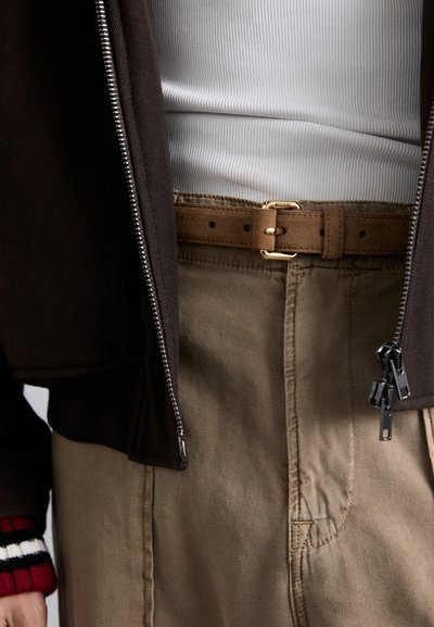 Pruun lukuga jakk, valge ribide mustriga tank top, khakivärvi püksid, pruun nahkrihm kuldse pandlaga ja punaste triipudega mansetid on nähtavad.