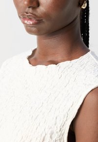 Haut sans manches texturé blanc avec un col en vagues, présentant un motif en relief et un tissu doux et léger.