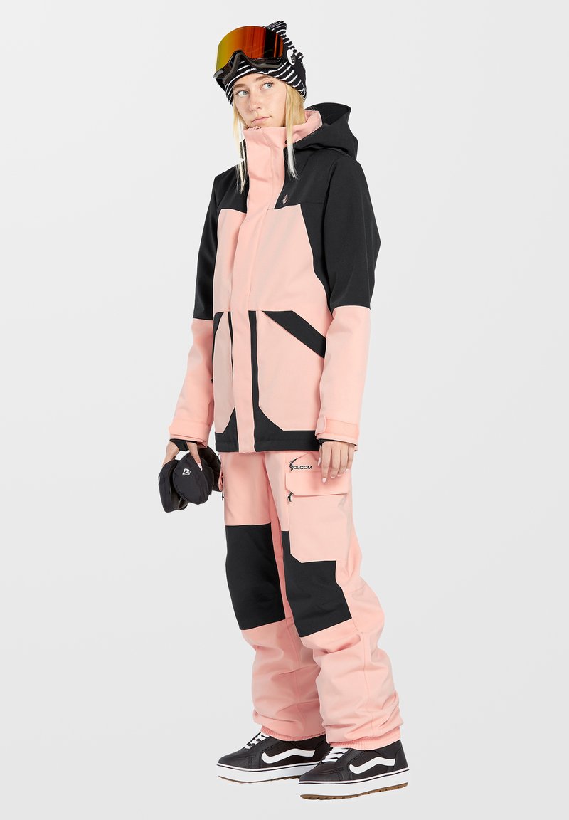 Roze en zwarte snowboardoutfit met een hoodie-jack en cargo-broek. De stof lijkt getextureerd, met een ontwerp in meerdere panelen en zakken.