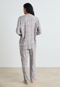 Femme debout pieds nus, de dos, portant un pyjama gris assorti avec des motifs abstraits en forme de corde blanche.