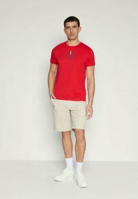 T-shirt de algodão vermelho com decote redondo e impressão gráfica. Combinado com calções bege, sapatilhas brancas e meias brancas. Fundo neutro.