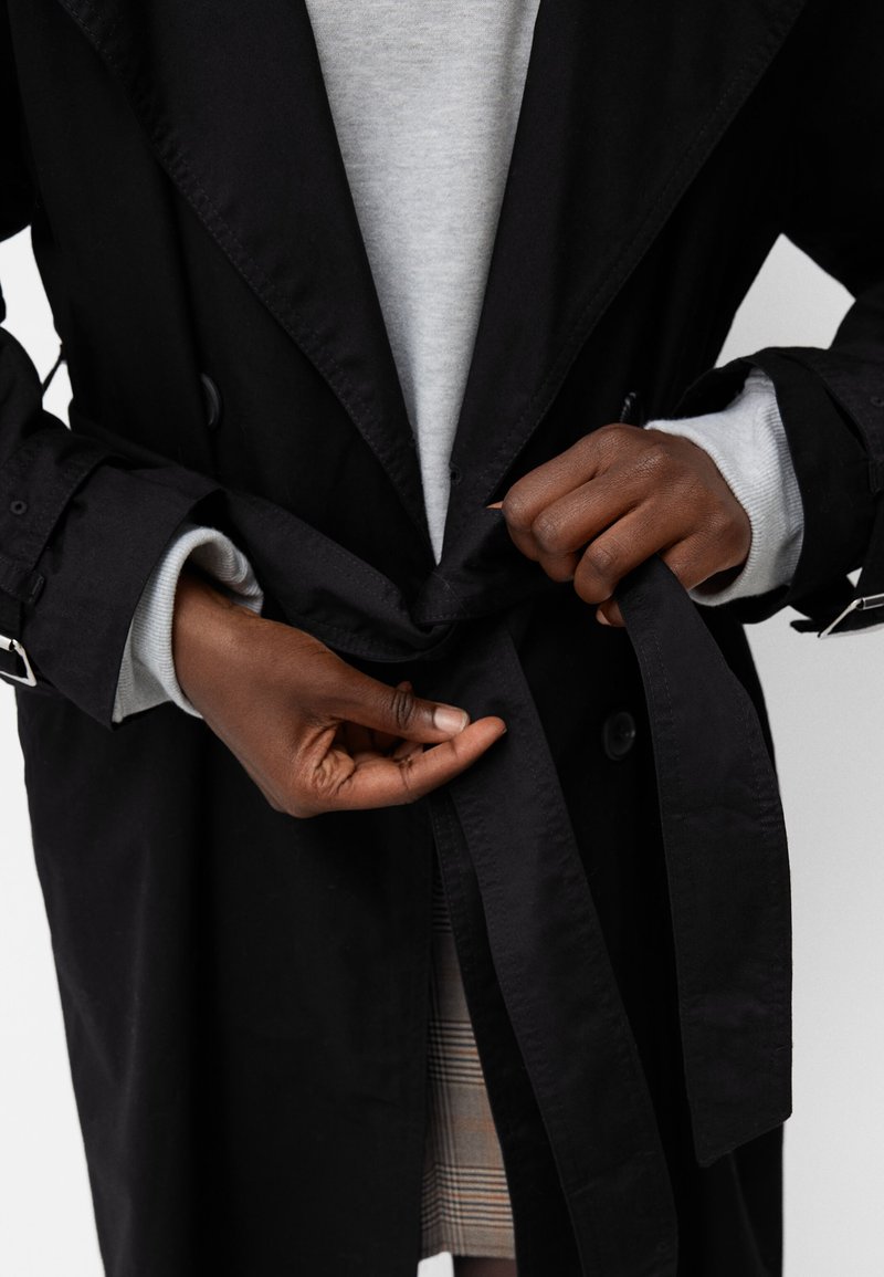 Czarny trencz z szerokim kołnierzem i paskiem w taliach, z detalami w postaci guzików. Ręce trzymające pasek, nałożone na szary sweter.