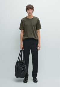 Olive green cotton t-shirt, black trousers, holding a large black leather bag with textured finish and double straps. Casual fit.