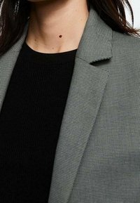 Grijze gestreepte blazer met een notch lapel, daaronder een gestructureerde zwarte top met een subtiele houndstoothpatroon en een gladde stofafwerking.