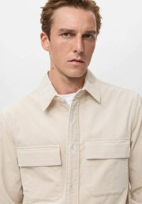 Camisa de pana beige con cuello puntiagudo, dos bolsillos frontales con tapa y cierre de botón. Tejido texturizado con rayas verticales.