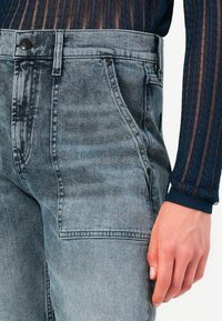 Gros plan sur une personne portant un pantalon en denim gris délavé à taille haute avec une grande poche avant, associé à un haut à manches longues texturé bleu marine.