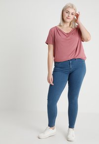 Pink draped top with short sleeves paired with blue skinny jeans and white sneakers, standing on a neutral background.