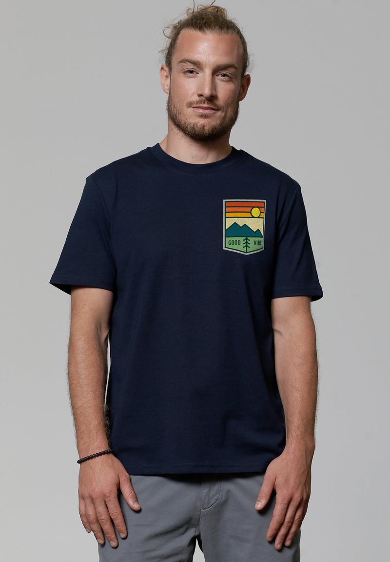 Dunkelblaue Baumwoll-T-Shirt mit einem bedruckten Patch, der Berge, Sonne und den Text "GOOD VIBE" in Orange und Grün zeigt. Kurze Ärmel.