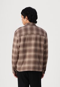 Camicia in flanella a quadri marrone con colletto, maniche lunghe e orlo dritto. Presenta un motivo a scacchi alternato tra chiaro e scuro.
