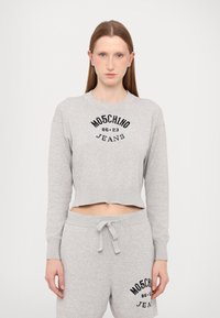 Sweatshirt cinzento cortado com punhos canelados, apresentando texto gráfico preto "MOSCHINO JEANS 86-23" no peito, combinado com calças de moletom a condizer.