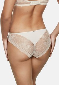 Sous-vêtements en dentelle beige avec un motif floral et des bords festonnés. Présente un panneau avant lisse et un dos en dentelle délicat pour une texture détaillée.