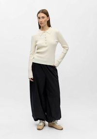 Ribbet, cremefarvet top med lange ærmer, krave og lukning med fem knapper, kombineret med løse, sorte bukser med vide ben og lysebrune sneakers.