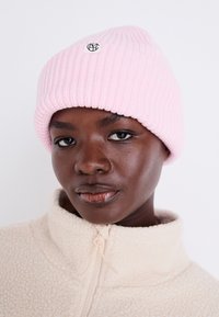 Rosa gerippte Mütze mit umgeschlagenem Saum und einem runden schwarzen Logo an der Vorderseite. Kombiniert mit einer beigen Fleecejacke mit hohem Kragen.