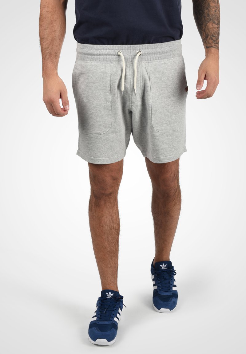 Helle graue Baumwollshorts mit einem elastischen Bund, verstellbarem Kordelzug und zwei Seitentaschen, kombiniert mit dunkelblauen Turnschuhen.