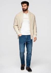 Cardigan beige en tricot porté sur un polo blanc avec un accent rayé, associé à un jean bleu et des chaussures marron foncé. Coupe décontractée et tissu texturé.