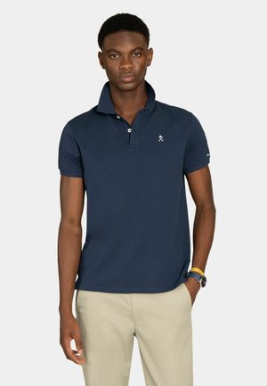 Joven con camisa polo azul marino con el cuello levantado, pantalones beige, reloj y pulsera, de pie con una mano en el bolsillo.