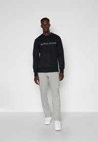 Μαύρη ζακέτα με λογότυπο, γκρι παντελόνι φόρμας και λευκά αθλητικά παπούτσια. Το ντύσιμο διαθέτει χαλαρή εφαρμογή και μαλακή υφή υφάσματος.